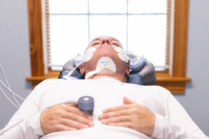 A male BeachGlo Aesthetics client lies on a treatment table, wearing the Emface's cheek and submentum applicators. Emface near Kitty Hawk tightens skin and tones muscle to improve facial structure.