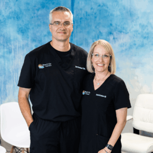 Co-founders Ben and Sarah Kershner stand smiling in their office, in front of a blue wall and plush white chairs. As experts in laser back hair removal in Nags Head, NC, Ben and Sarah help every patient achieve their aesthetic goals.